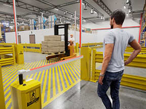 Pedestrian waiting behind yellow AisleCop gates while a loaded forklift crosses a four-way intersection