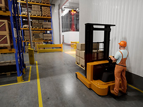 Walk-behind stacker operator approaching a crossing with an overhead warning light mounted at the far end of the aisle